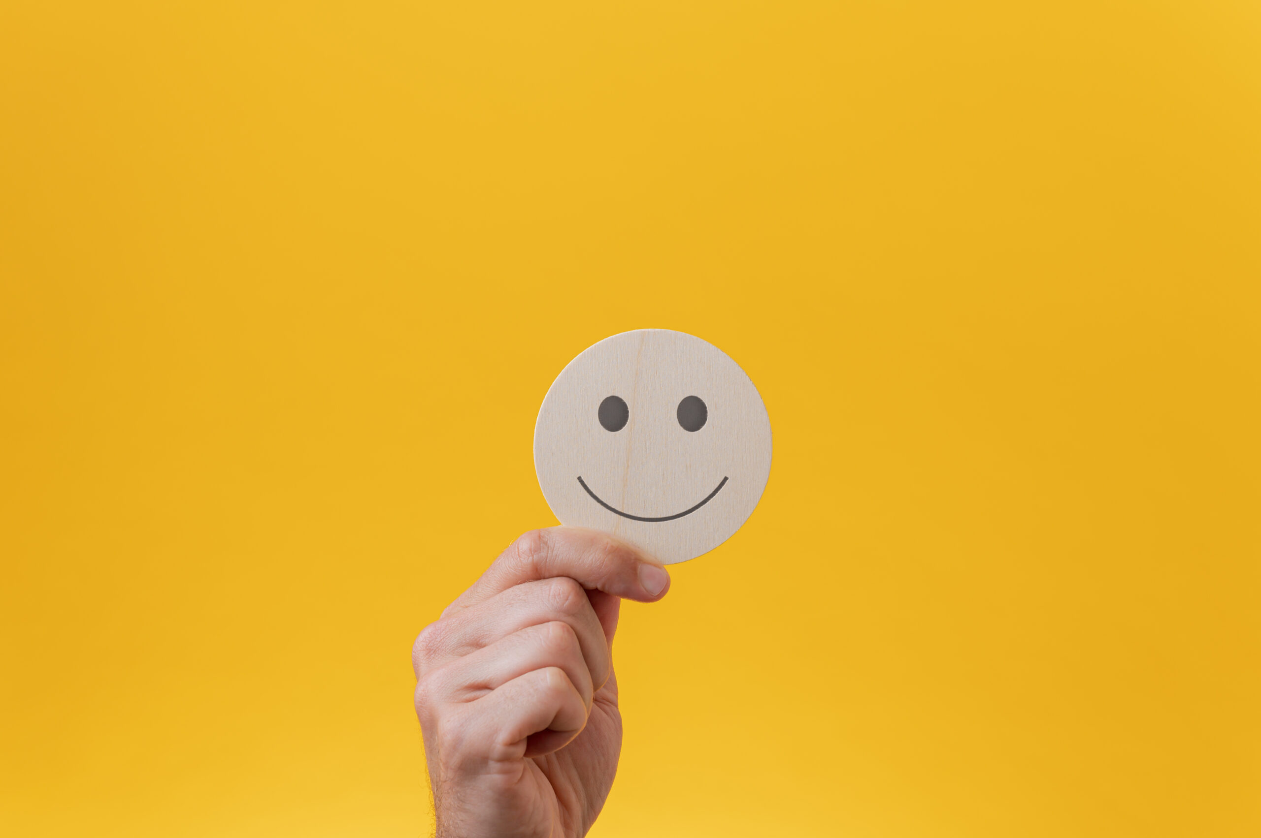 Mano maschile che mostra un cerchio tagliato in legno con sopra un volto sorridente. Su sfondo giallo.