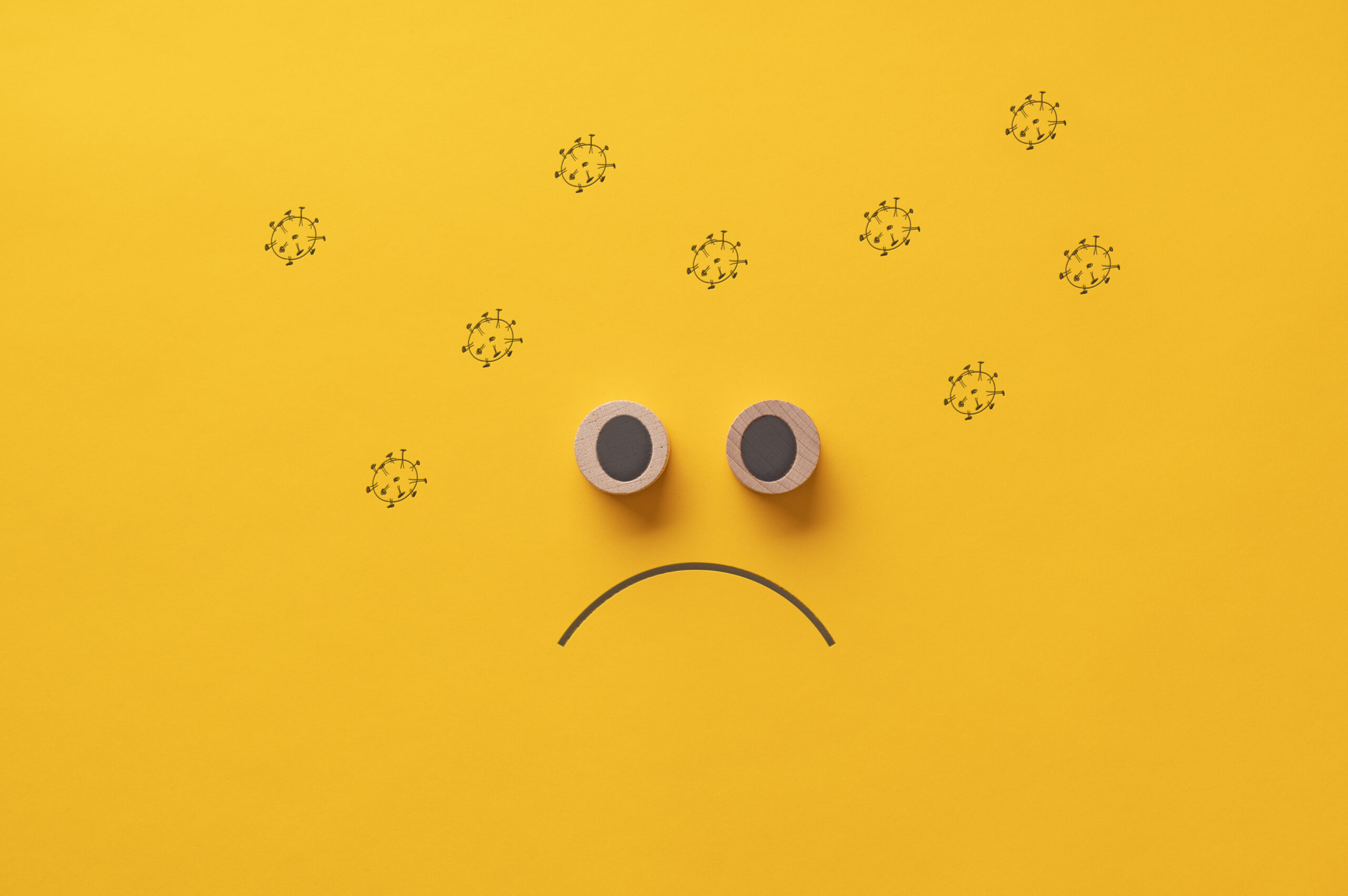 Sad face made of wooden cut circles surrounded with hand drawn coronavirus molecules. Over yellow background.
