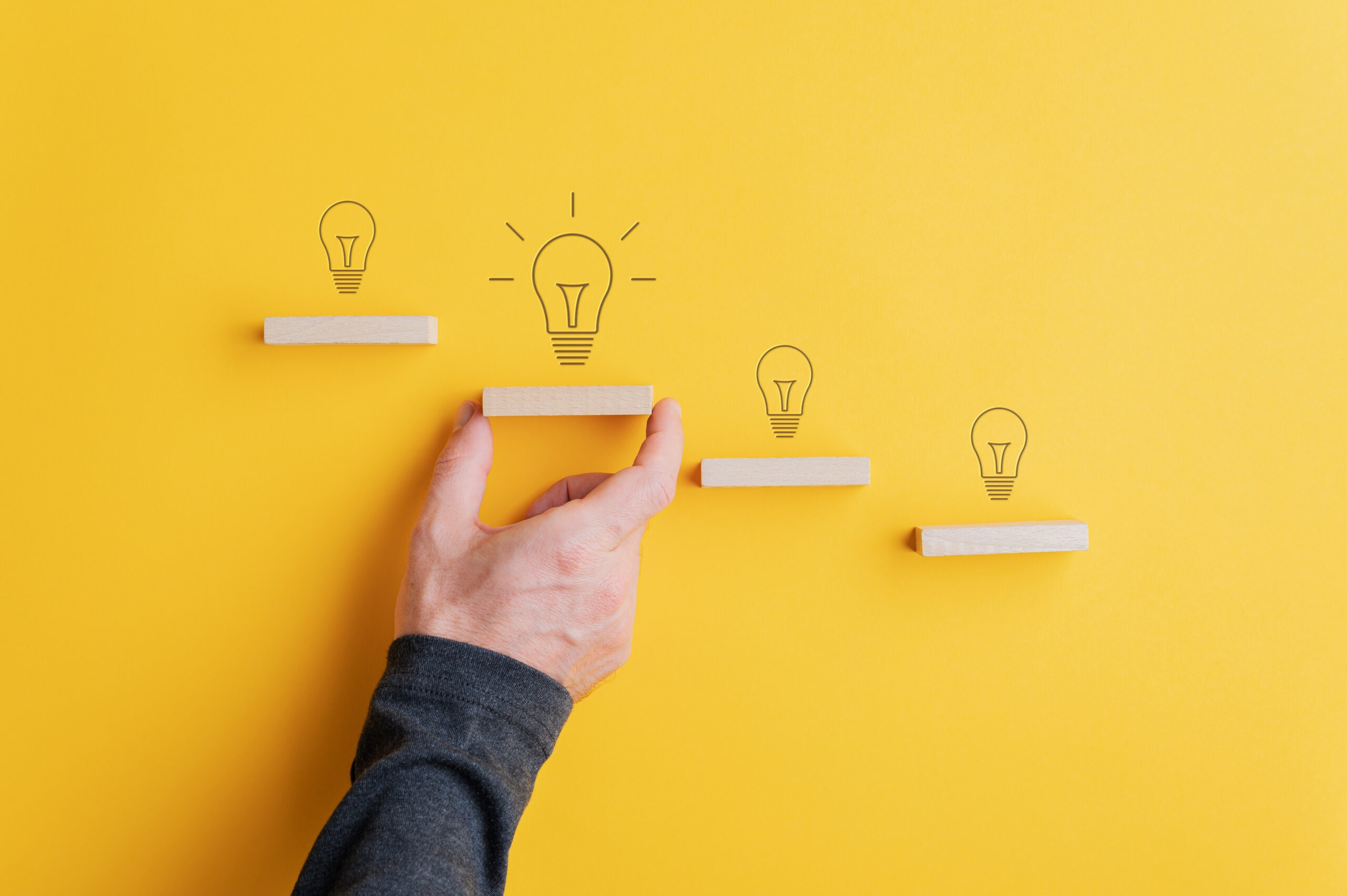 Light bulbs drawn above each step in conceptual stairway of wooden pegs with male hand supporting the one with the biggest and glowing bulb.